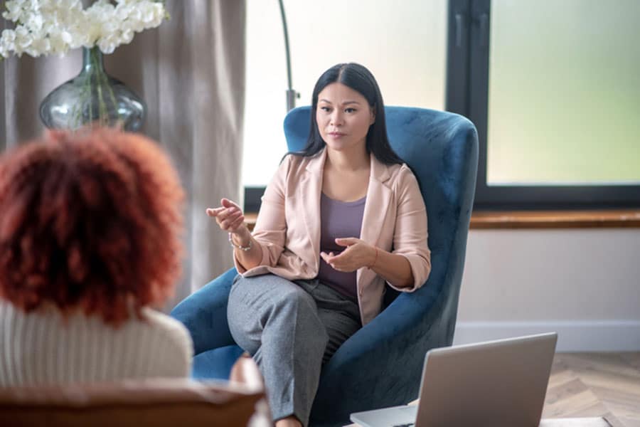 A woman sits in a blue chair, gesturing while talking to another person. A laptop is open on a table nearby, and a window and flowers are visible in the background.